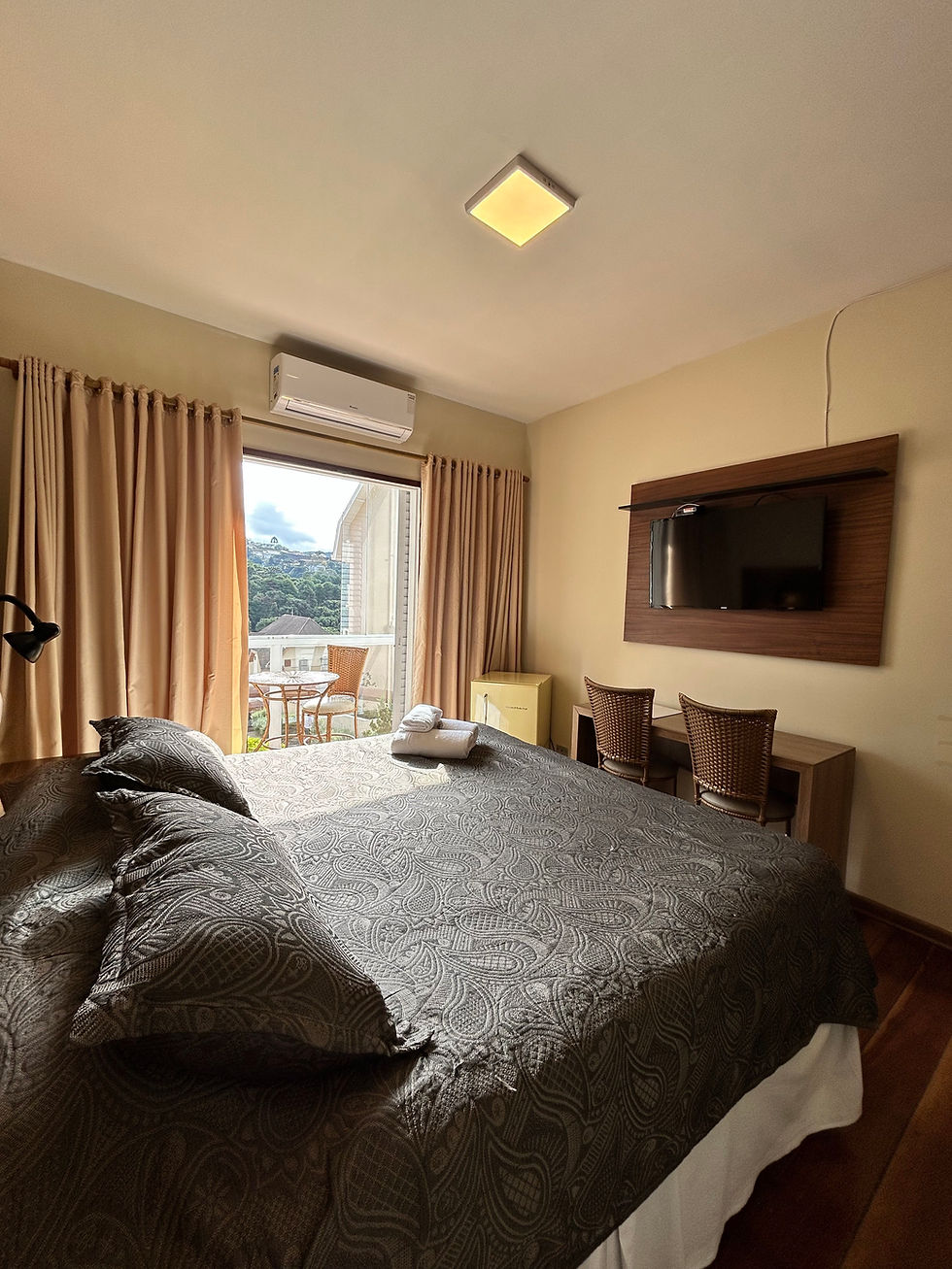 Vista da suíte da Pousada Kaliman com cama de casal, televisão, frigobar e varanda com vista para Campos do Jordão