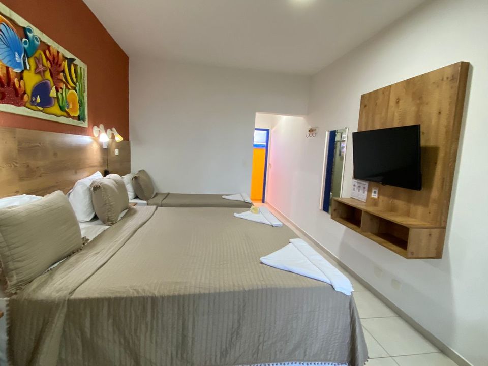 Suíte com cama de casal, cama de solteiro, televisão de tela plana e pintura colorida na parede cor de ferrugem. Porta azul para banheiro iluminado em amarelo, paredes brancas e piso de cerâmica branca. Colchas bege listradas e toalhas brancas sobre as camas em Ubatuba na Pousada Kaliman