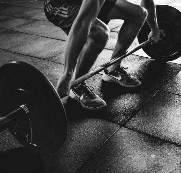 A black and white photo of an athlete squatting to lift a barbell on the gym floor. The athlete is wearing shorts and sneakers, at Trein Club in Houston