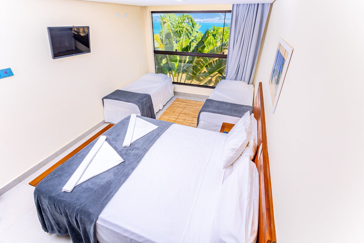 Suíte do Eco Baepi com uma cama de casal e duas camas de solteiro e janela com vista para o mar e serra em Ilhabela