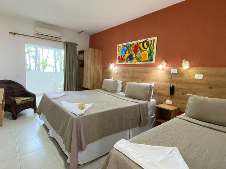 Quarto de hotel aconchegante com cama de casal, solteiro, parede laranja-terra, arte marinha, ar condicionado e roupas de cama claras com toalhas brancas. Possui cadeira de vime estampada, janela com vista e guarda-roupa de madeira clara em Ubatuba na Pousada Kaliman
