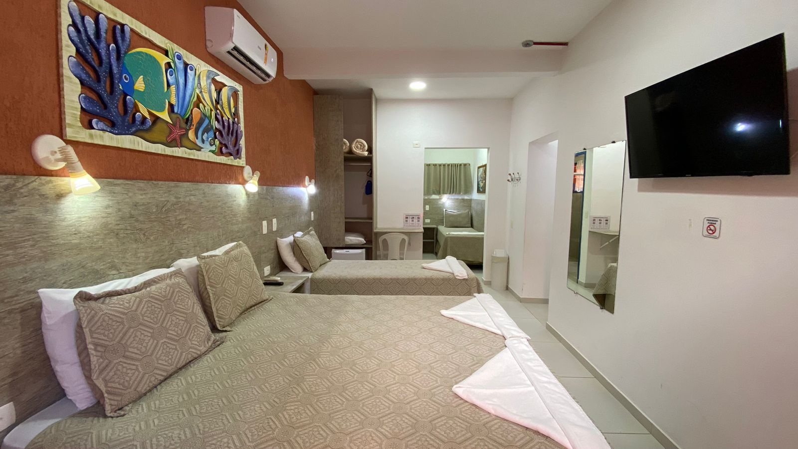 Suíte da Pousada Kaliman com cama de casal e solteiro, edredons bege e travesseiros cinza, com toalhas brancas enroladas na cama de casal. A parede ferrugem atrás da cama tem cabeceira de madeira clara, pintura marinha e arandelas brancas. O quarto tem paredes brancas, piso bege e porta laranja com detalhes azuis ao lado da mesa de cabeceira na praia de Ubatuba