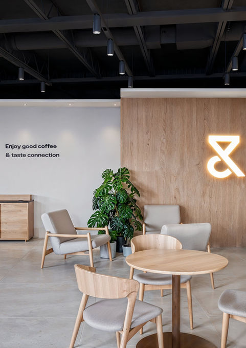Modern, well-lit café. In the center, there is a round wooden table with four curved-back wooden chairs with gray upholstery. In the background on the right, a wooden wall features a stylized orange neon Trein Club logo