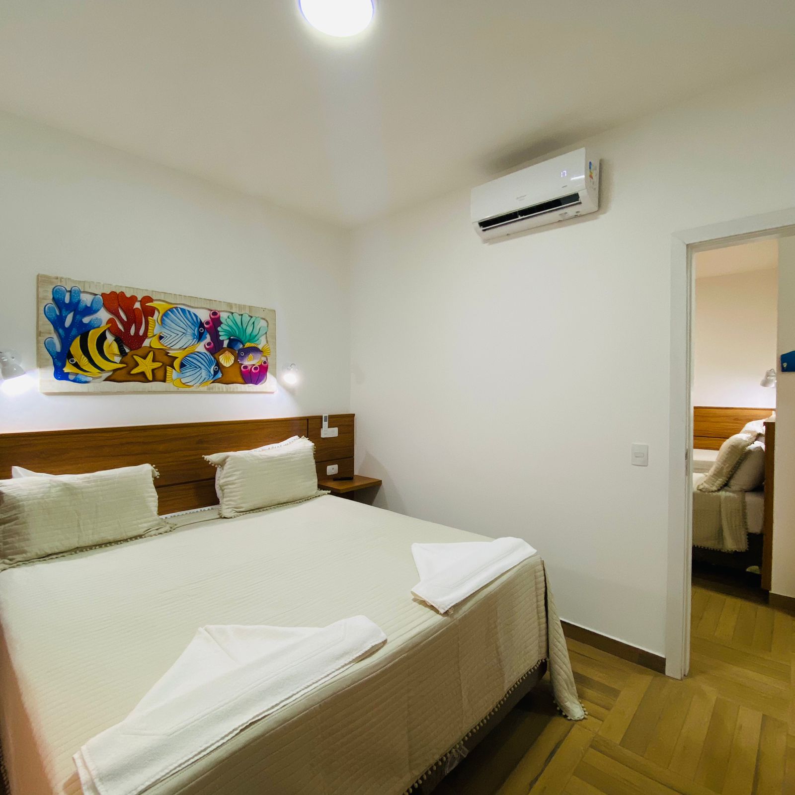 Suíte da Pousada Kaliman com cama de casal, cabeceira de madeira e roupa de cama clara. Toalhas brancas dobradas sobre a cama. Uma pintura com tema marinho decora a parede. Há ar-condicionado e uma porta aberta que revela outro quarto com mais uma cama. Piso de madeira clara na cidade de Ubatuba