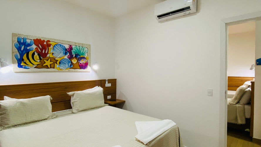 Quarto de hotel em Ubatuba com cama de lençóis brancos, obra de arte marinha colorida acima da cabeceira de madeira e ar condicionado na parede. Porta aberta à direita para outro quarto e paredes brancas na Pousada Kaliman