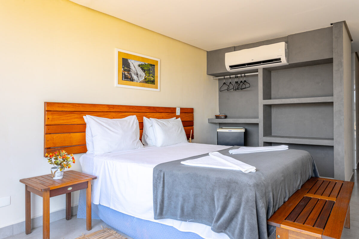 A suíte Eco Baepi Residence conta com uma cama de casal posicionada em frente a uma televisão, parede amarela é adornada com um quadro, e o ambiente conta com um pequeno frigobar, acima espaço para roupas com alguns cabides disponíveis