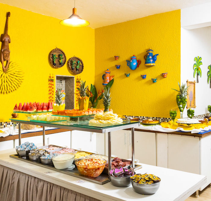 Buffet de café da manhã vibrante da Pousada Kaliman em Ubatuba com decoração tropical e paredes amarelas decoradas. Mesa longa com frutas frescas (melancia, mamão, abacaxi), queijos, presuntos e outros itens matinais, além de cereais e iogurtes. Ao fundo, outra mesa com opções. Iluminação brilhante realça as cores vivas.