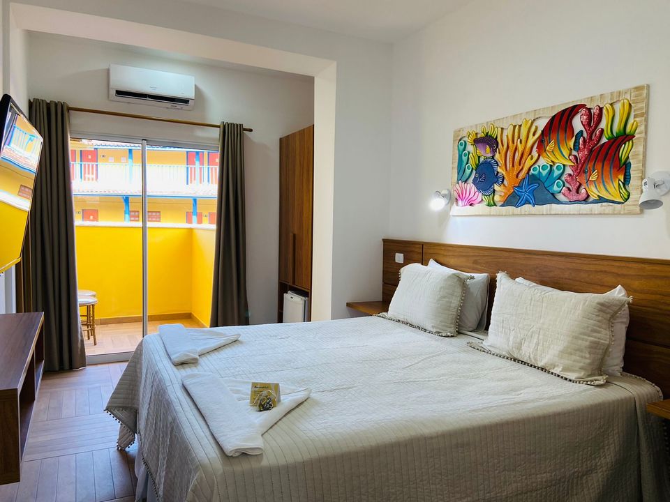 Acomodação da Pousada Kaliman com cama de casal decorada com cisne de pétalas vermelhas, pintura marinha em parede laranja. Porta de vidro para varanda amarela com vista para palmeiras e céu azul. Ar-condicionado branco. Canto com mesa redonda, toalha vermelha, duas taças e pétalas no chão de cerâmica clara na praia de Ubatuba
