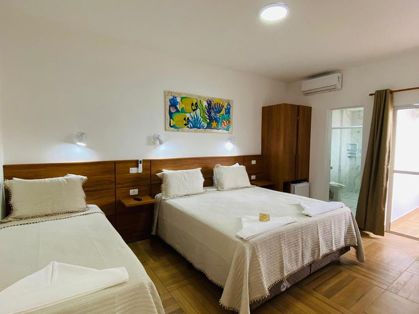 Quarto de hotel iluminado e organizado na praia de Ubatuba com uma cama de casal, uma de solteiro, cabeceiras de madeira com luminárias e prateleiras. Destaque para pintura colorida de peixes sobre a cama de casal. Guarda-roupa escuro, frigobar, porta para o banheiro e janela com cortinas marrons claras completam o ambiente na Pousada kaliman
