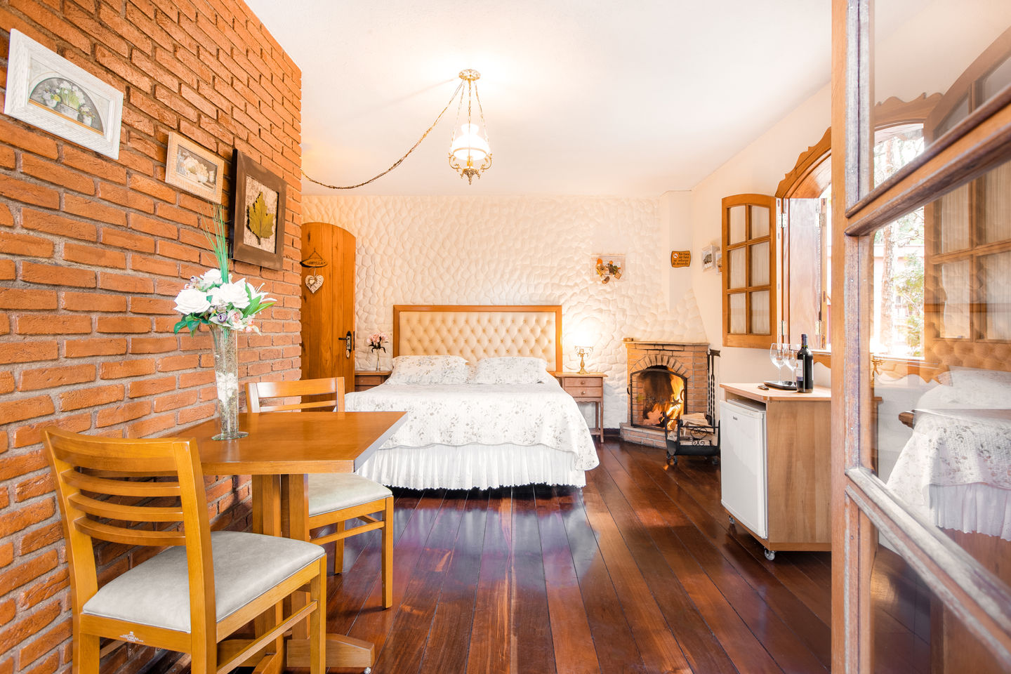 Quarto com parede de tijolos e gesso, cama com cabeceira de couro bege, lareira acesa, mesa com flores e frigobar embutido na Estalagem Spiller em Monte Verde
