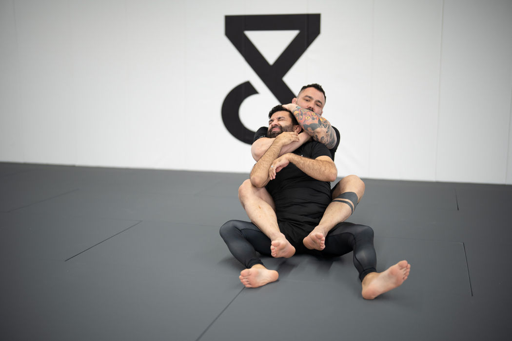 Two men practicing jiu-jitsu on a gray mat at Trein Club, with a white wall displaying a large logo in Houston