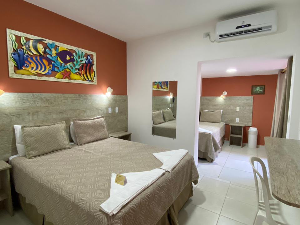 Suíte em Ubatuba com cama de casal, cama de solteiro em outro ambiente e pintura marinha colorida na parede cor de ferrugem. Paredes brancas, cabeceira cinza texturizada, espelho e ar-condicionado branco. Porta aberta para o outro quarto com cama de solteiro e piso de cerâmica clara na Pousada Kaliman
