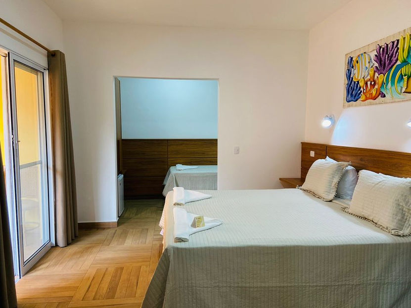 Quarto de hotel amplo e iluminado na Pousada Kaliman com cama de casal (edredom branco, cabeceira de madeira clara, abajur), pintura abstrata azul, laranja e amarela, porta de vidro com cortinas marrons (acesso à varanda ensolarada) e outro ambiente ao fundo com cama de solteiro e móveis de madeira clara em Ubatuba
