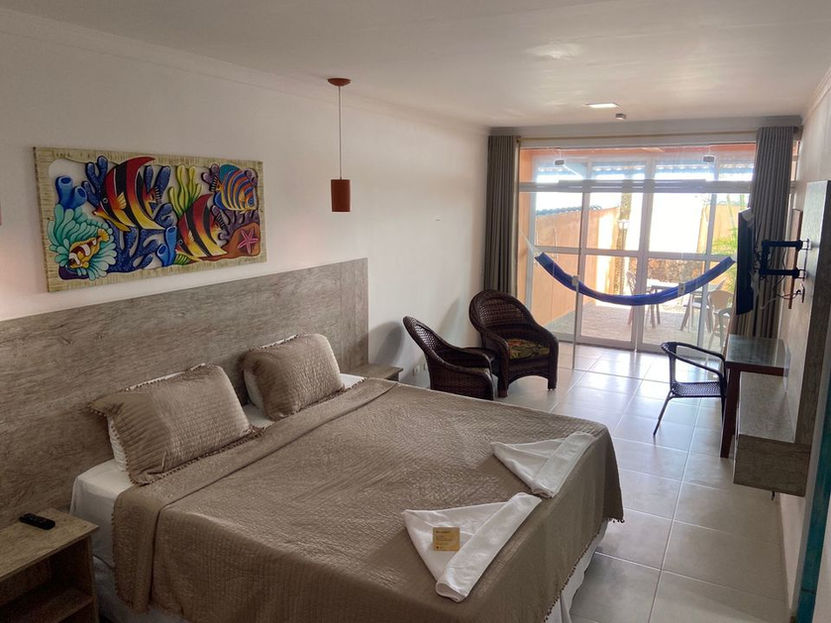 Quarto de hotel bem iluminado com cama grande, lençóis bege e travesseiros brancos. Decoração inclui obra de arte colorida com peixes e corais acima da cama, duas cadeiras de vime escuras e mesa com TV de tela plana. Janelas do chão ao teto dão para pátio com rede azul e mobiliário de vime na Pousada Kaliman em Ubatuba