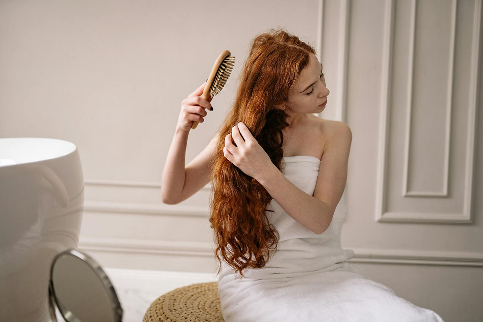 Woman in a white towel brushes long red hair in a minimalist bathroom. Neutral-toned walls, a round wicker seat, and a small mirror visible.