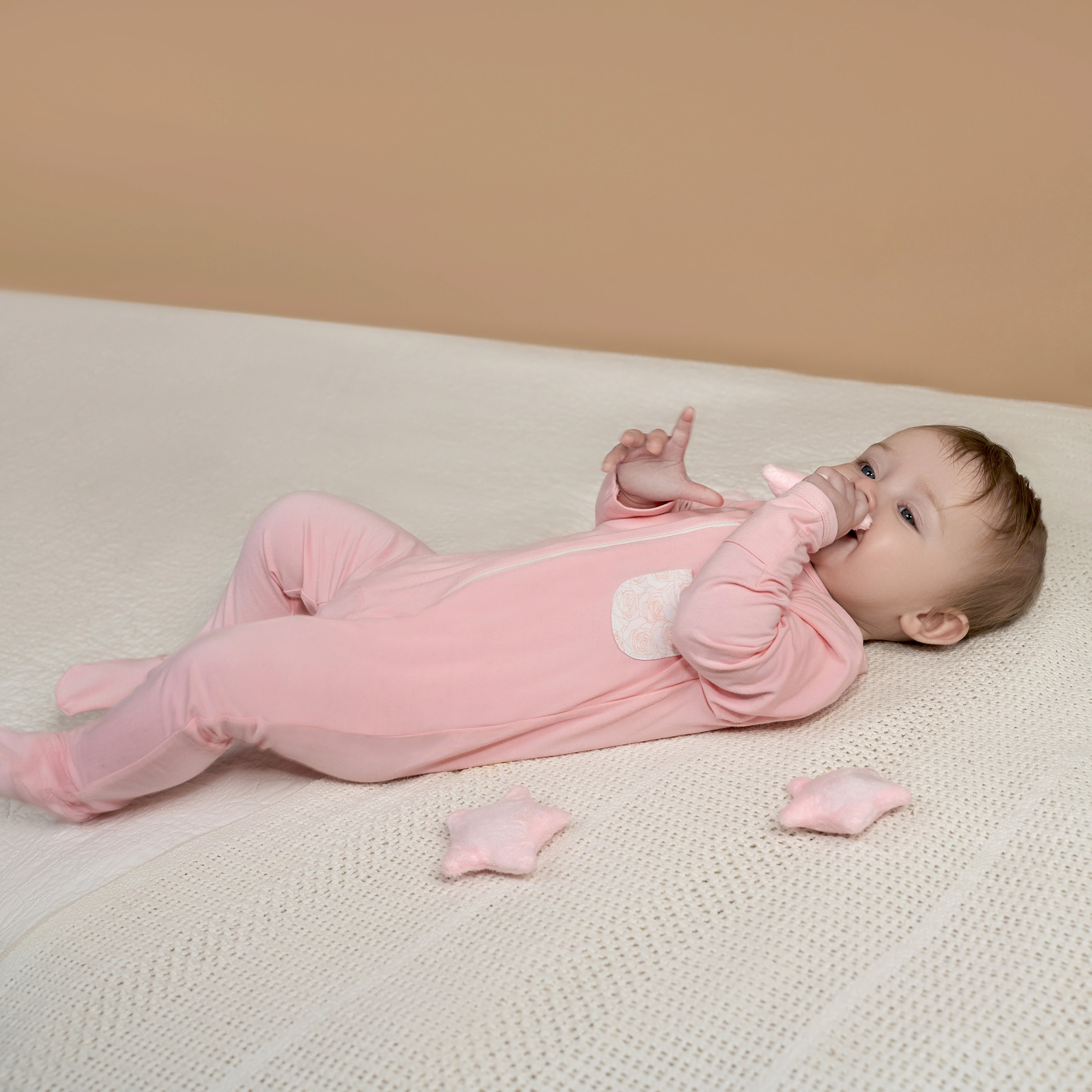 Cheerful baby in pink sleepsuit lying on a cream blanket, reaching out to play with star-shaped toys.