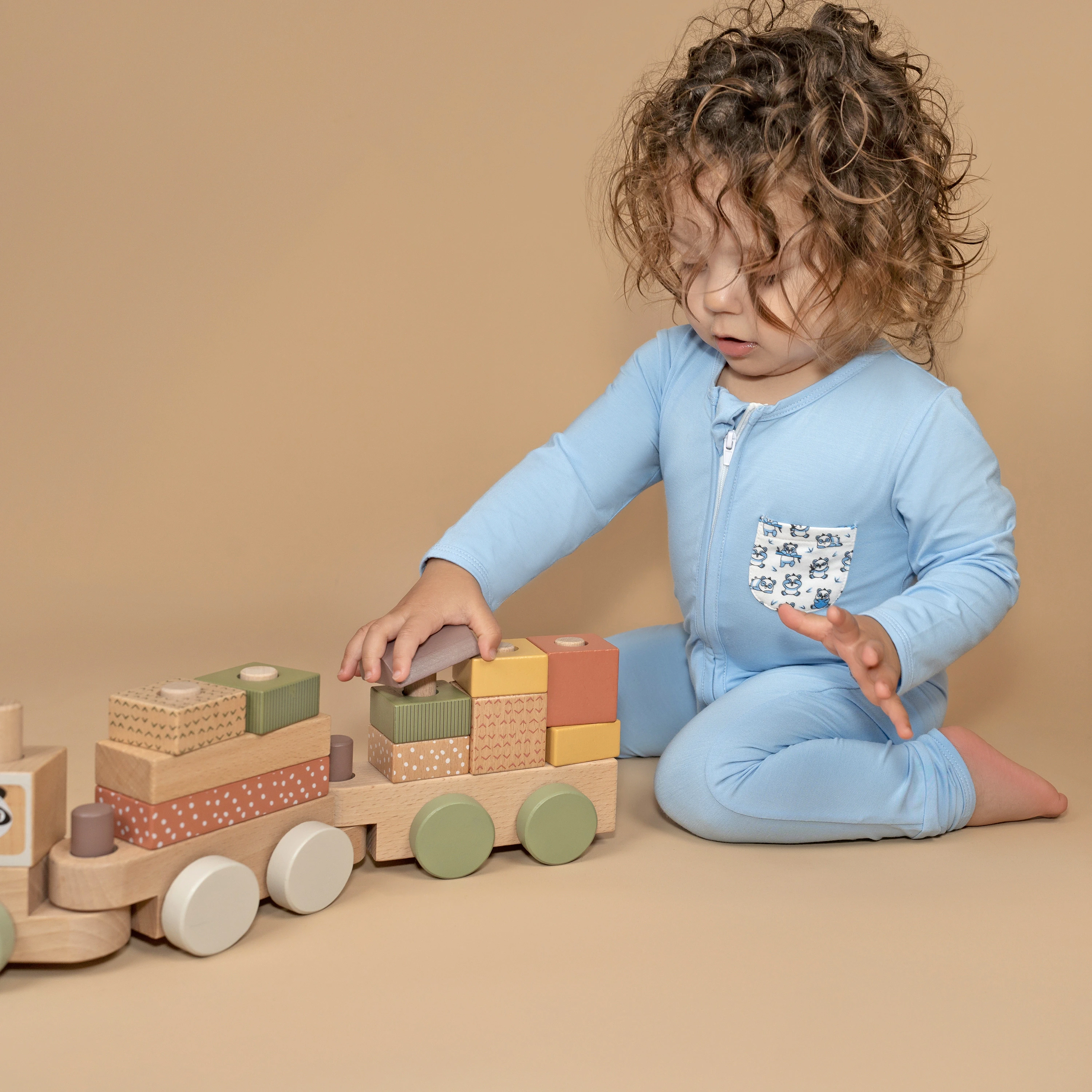 Thoughtful toddler in a light blue sleepsuit featuring pandas, engaged with wooden toys on a soothing beige surface.