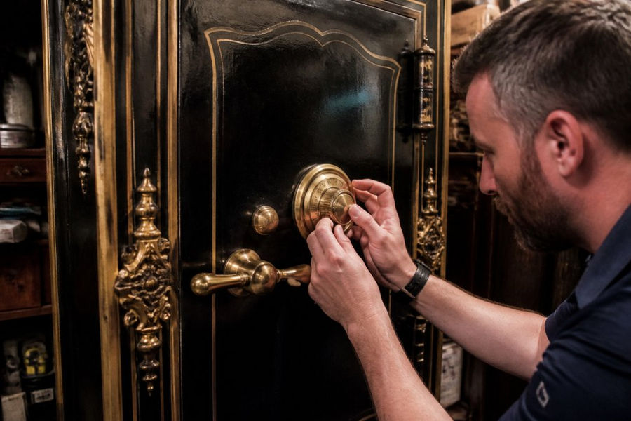 A locksmith's hands carefully turning the dial on a large, ornate antique safe with brass fittings in a dimly lit room, showing the intricate details of the safe's door and locking mechanism