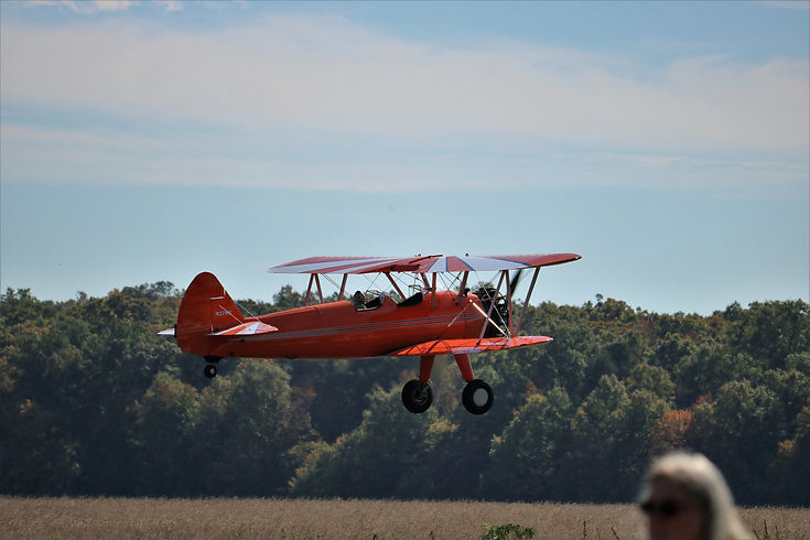 Red biplane flying above a tree-lined airfield