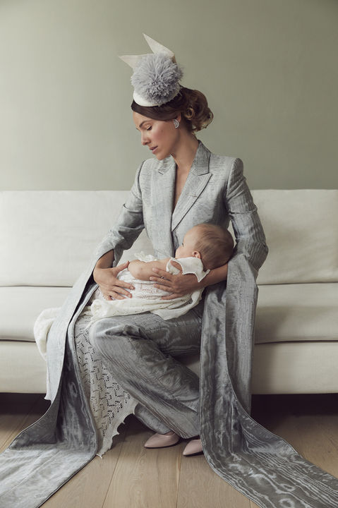 Portrait inspired by British monarch aesthetics, featuring actress Romi Aboulafia and her newborn baby. Romi is dressed in an elegant silver suit with dramatic flowing details and a statement fascinator, cradling her baby wrapped in a delicate knit blanket. The composition exudes sophistication and maternal warmth, evoking timeless royal elegance.