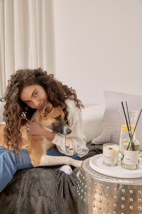 Shlomit Malka, international model, cuddling a dog on a cozy bed in a bright and elegant home setting, styled for a Castro Home campaign. A sleek metallic side table features a scented candle and diffuser, adding a touch of warmth and sophistication. Photography by Liron Weissman photographer