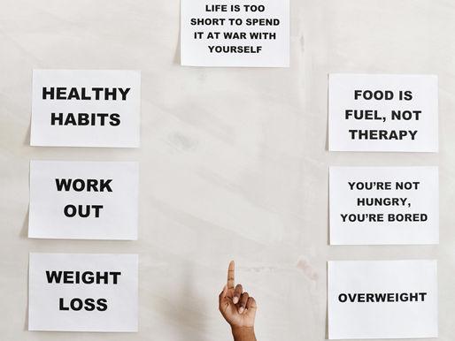 Imagen con mensajes motivacionales impresos en carteles blancos con letras negras sobre una pared clara. Los textos dicen en inglés: "Life is too short to spend it at war with yourself", "Healthy habits", "Work out", "Weight loss", "Food is fuel, not therapy" y "You're not hungry, you're bored". Se observa una mano apuntando hacia arriba, señalando uno de los carteles.
