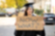 Sad Graduate Student Standing With Now What Placard On Street.jpg
