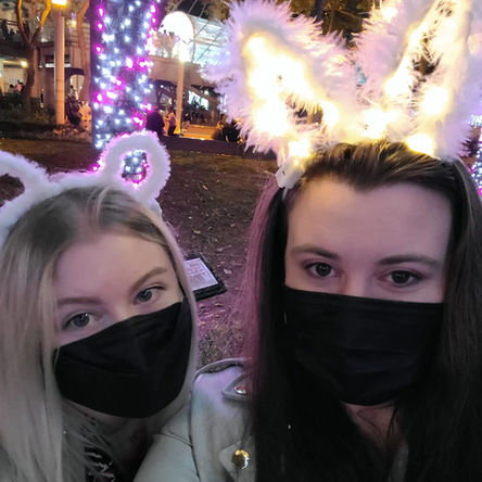 Two girls in black face masks wearing light up bunny and teddy bear eats taking a selfie in front of a Christmas tree wrapped in Christmas lights.