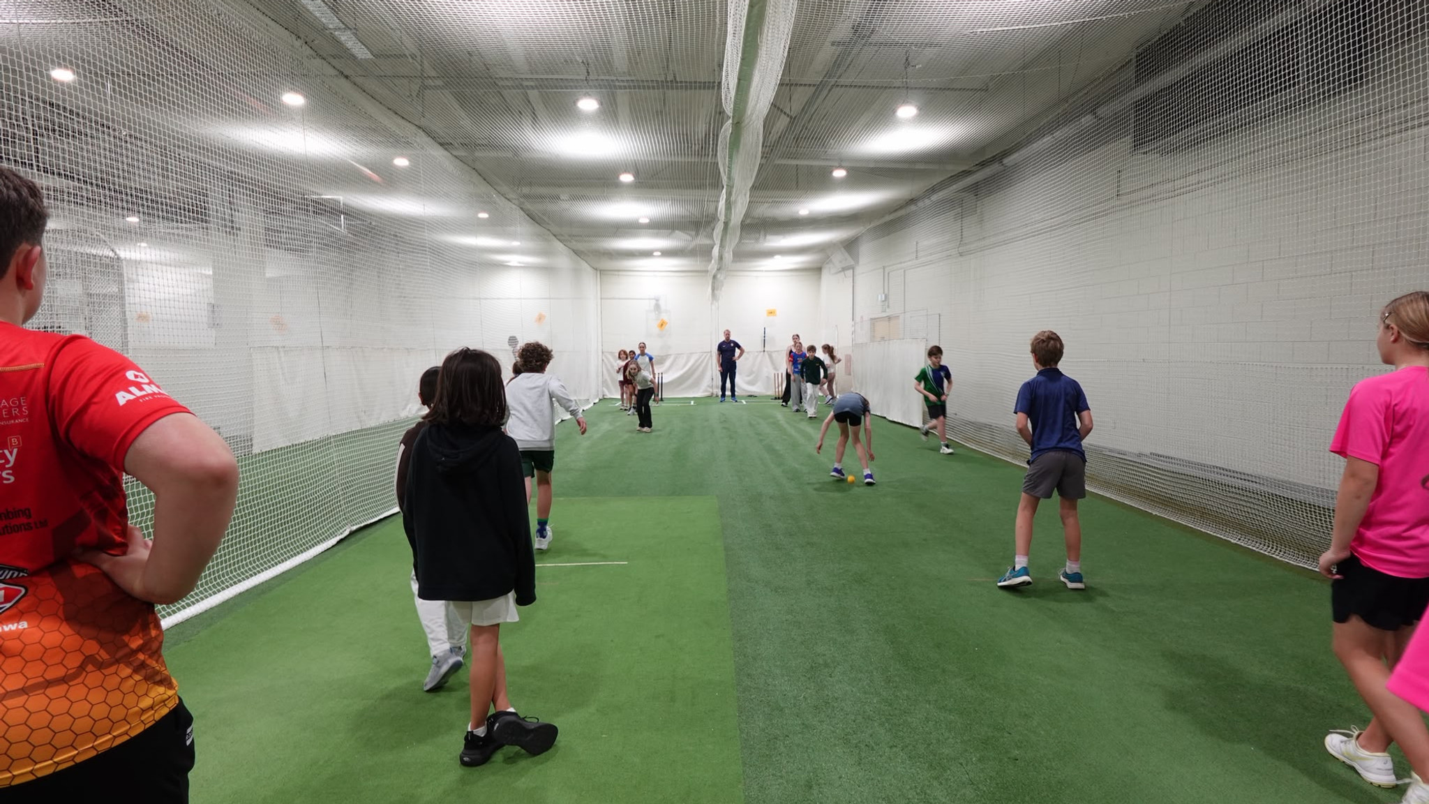 Kids practice cricket in an indoor netted training lane, with coaches watching on green turf under bright lights.