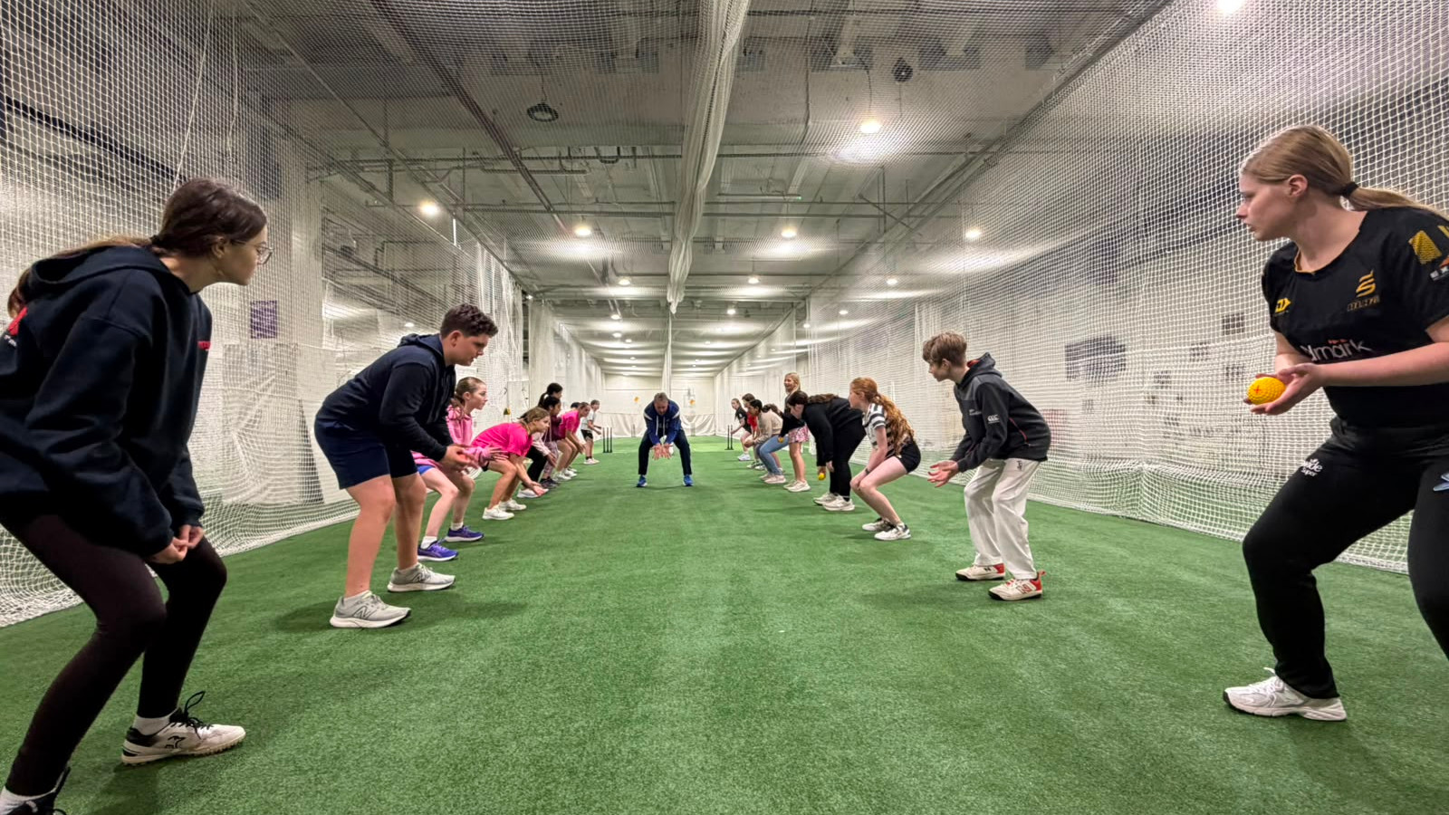 Kids crouch in a netted indoor turf lane as a coach drills fielding with a yellow ball; focused training session.