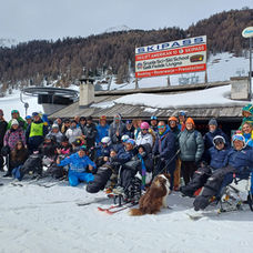 09:35Un gruppo di sciatori, alcuni con attrezzatura adattiva, sorridono su una pista innevata vicino a una seggiovia. Un edificio in legno espone un'insegna con la scritta "SKIPASS"