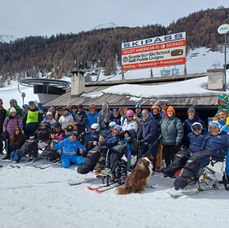 09:35Un gruppo di sciatori, alcuni con attrezzatura adattiva, sorridono su una pista innevata vicino a una seggiovia. Un edificio in legno espone un'insegna con la scritta "SKIPASS"