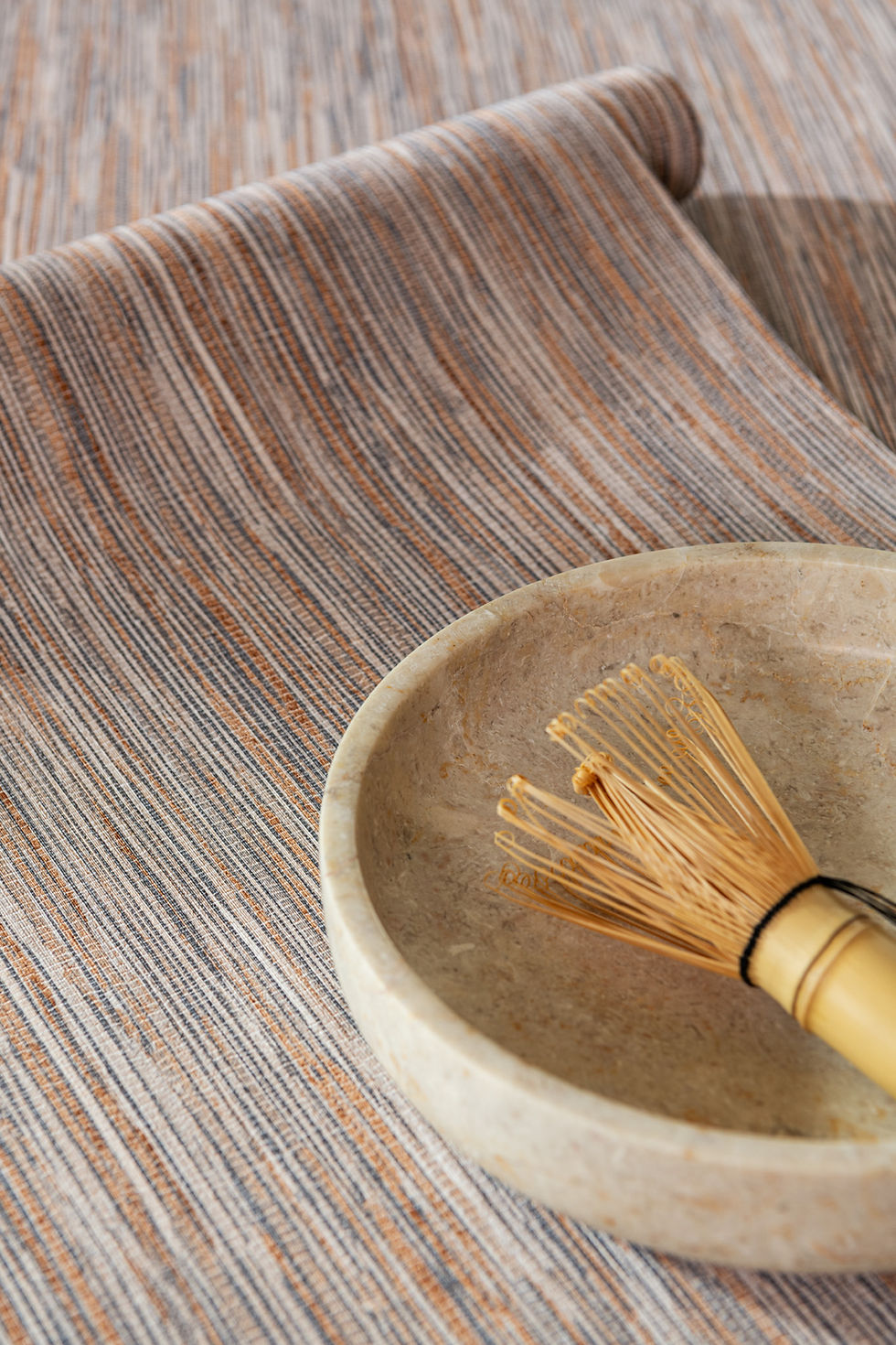 Ceramic bowl with a bamboo whisk on a brown-and-gray striped cloth, a calm minimalist tea-prep scene