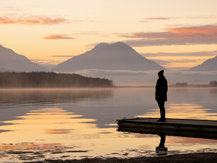 Person stands on a dock at sunset, overlooking a serene lake with misty mountains. Warm orange and pink hues fill the sky.