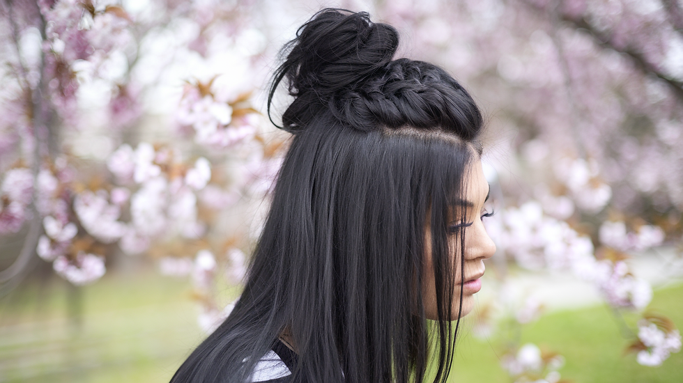 Woman with braided black hairstyle stands in profile among blooming pink cherry blossoms. Soft focus background, serene mood.