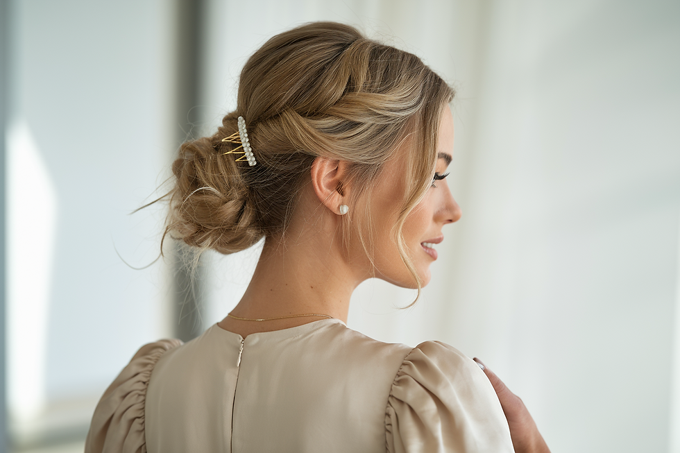Woman in a beige dress with elegant braided updo and pearl hairpin, smiling in bright room. Soft lighting enhances her calm demeanor.