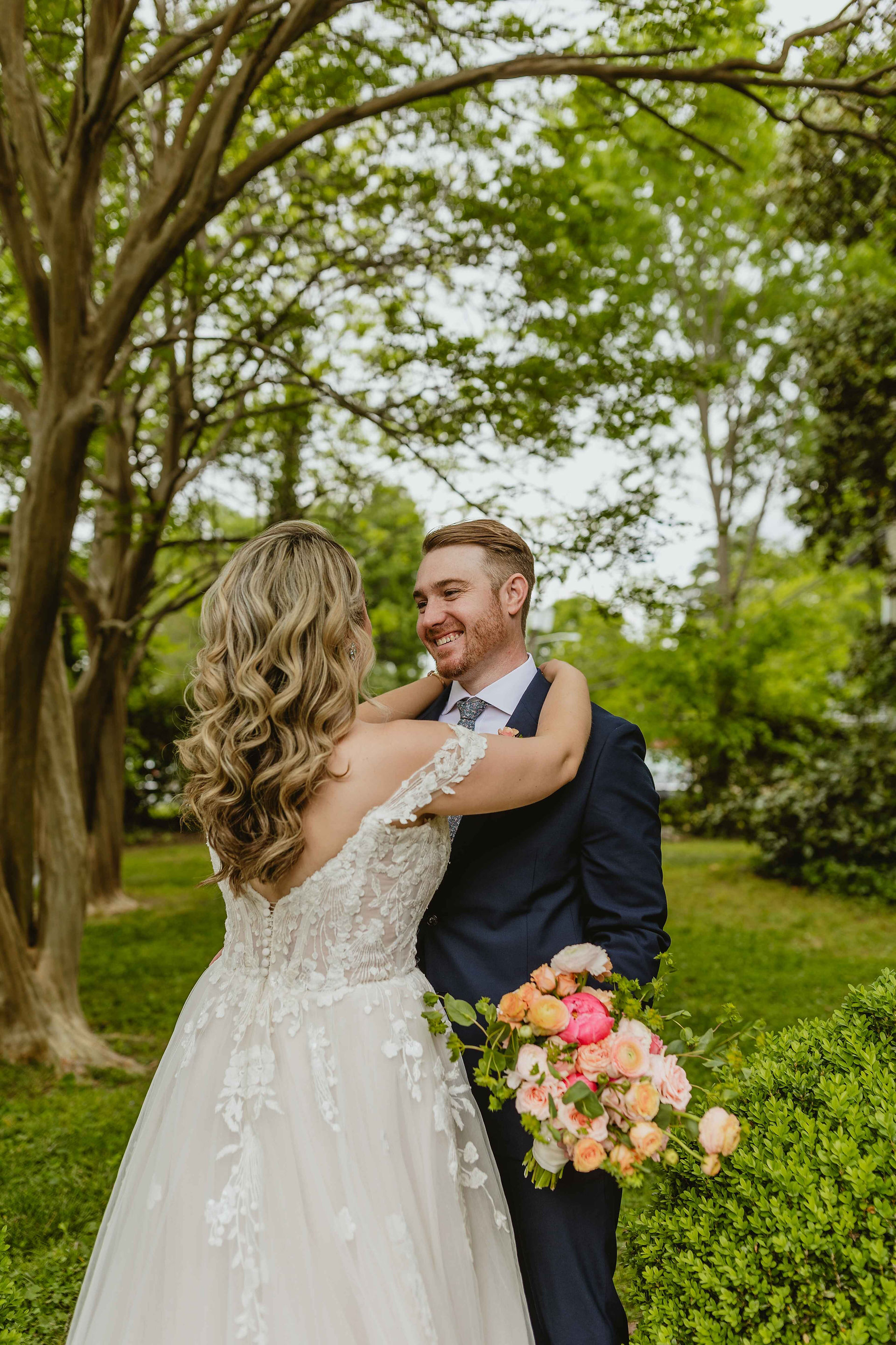 Groom is smiling at the bride holding her flowers bride is facing away. They are in the backyard of the Belk Mansion