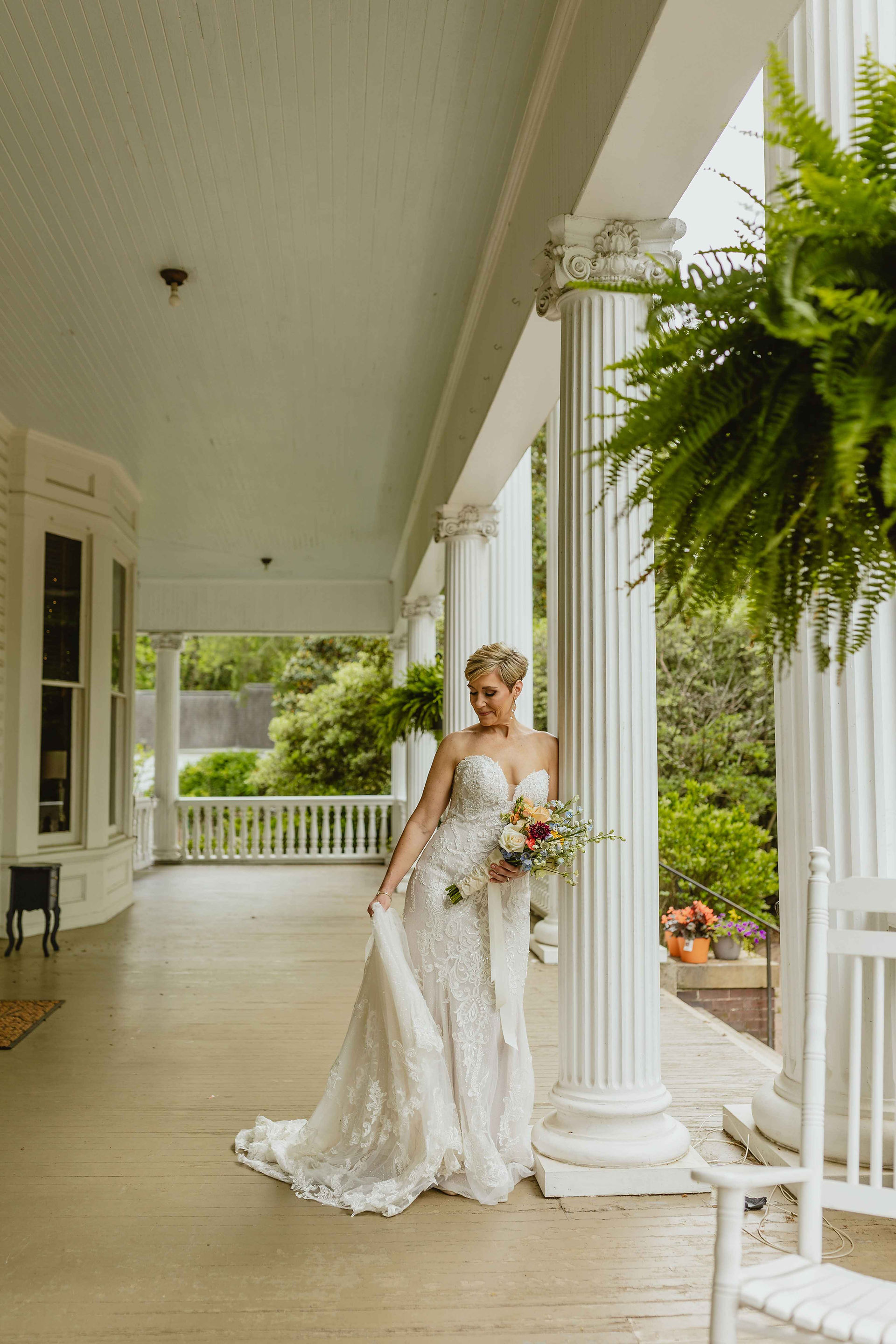 Bride is on the front porch holding her dress train. Looking down and holding her flowers in the other hand.