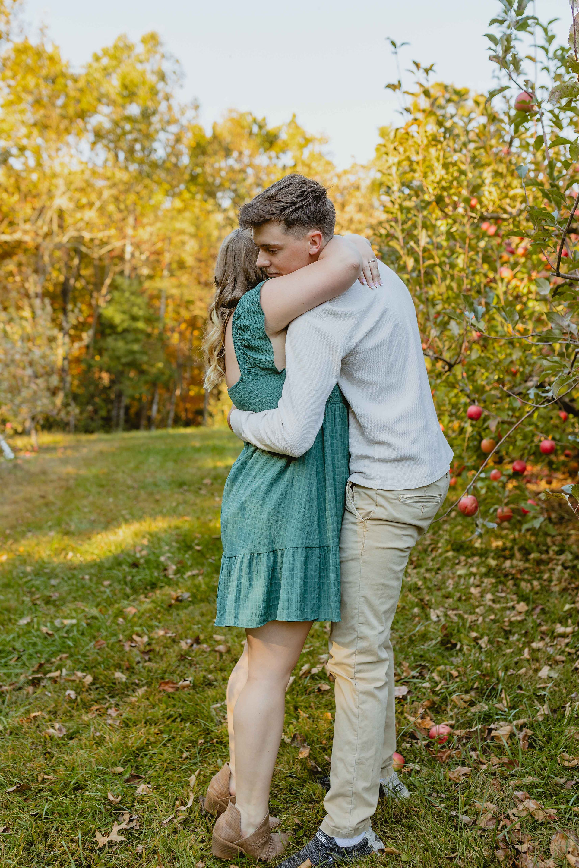 Couple is hugging each other after he proposed. There are apple trees and fall foliage behind them. 