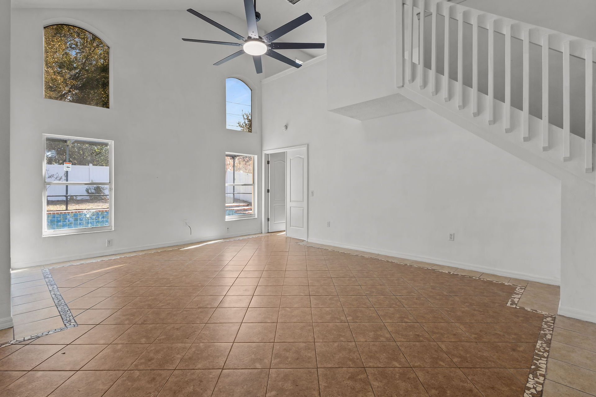 Empty living room with white walls, black ceiling fan, and stairs leading up to the loft