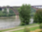 A bridge spans a river surrounded by lush green trees. The foreground features a park with a path, a lamppost, and manicured lawns.