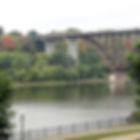 Bridge over a calm river with autumn trees in the background. A fence and lamp post are in the foreground, creating a peaceful scene.