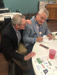Two men at a table, one leaning in with hand on the other's shoulder