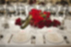 Elegant table setting with red roses centerpiece, white tablecloth, menus, candles, and black chairs. Romantic and sophisticated atmosphere.