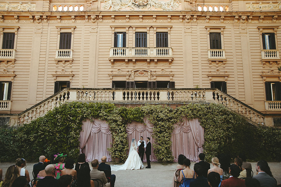 Bride and groom stand under textile draped backdrop for outdoor wedding ceremony at ornate building. Guests seated, surrounded by greenery. Elegant mood.
