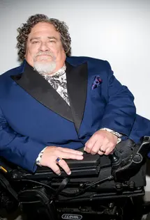 Jim LeBrecht in a dark blue Gucci suit with wide black satin lapels. A black and white floral silk shirt peeks out under the suit, and a dark blue handkerchief with red polka dots shows from the left suit pocket. He has dark gray curly hair, a salt and pepper mustache, and white beard and leans on his folded motor wheelchair.