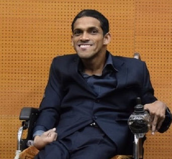 Akshansh Gupta with short dark hair and wearing a dark blue suit and dress shirt. He sits in a wheelchair and smiles at the camera.