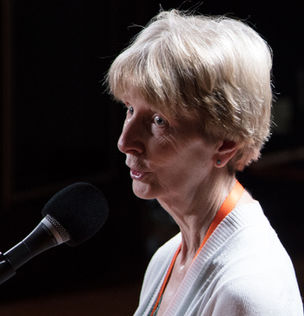Moe Keller speaks into a microphone. She has short white hair and bangs and wears a white sweater and orange lanyard around her neck.