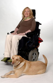 Barbara Turnbull sits in a powered wheelchair facing right, with a yellow Labrador retriever. She wears a brown shirt, khaki pants, green shoes, and a green and brown decorative neck scarf. She has curly blonde hair.