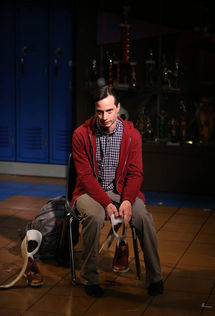 Gregg Mozgala in a red hoodie sits in a chair on stage holding a lower leg brace. A backpack and additional leg brace appear next to his chair, and the stage scenery includes a row of lockers and a glass case full of trophies.